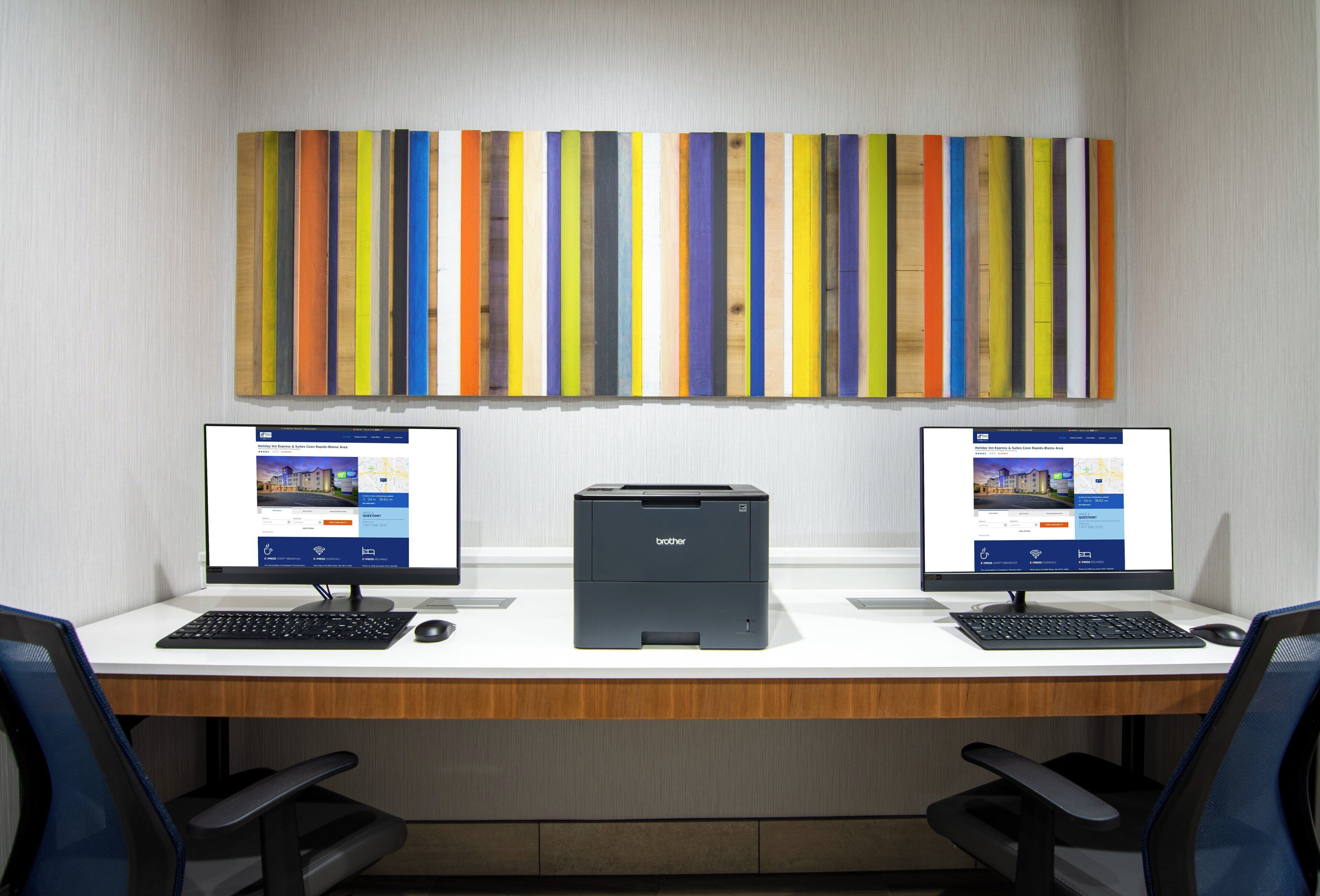 Un espacio de trabajo con dos computadoras de escritorio, una impresora y una obra de arte vertical de colores en la pared.