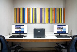 Un espacio de trabajo con dos computadoras de escritorio, una impresora y una obra de arte vertical de colores en la pared.