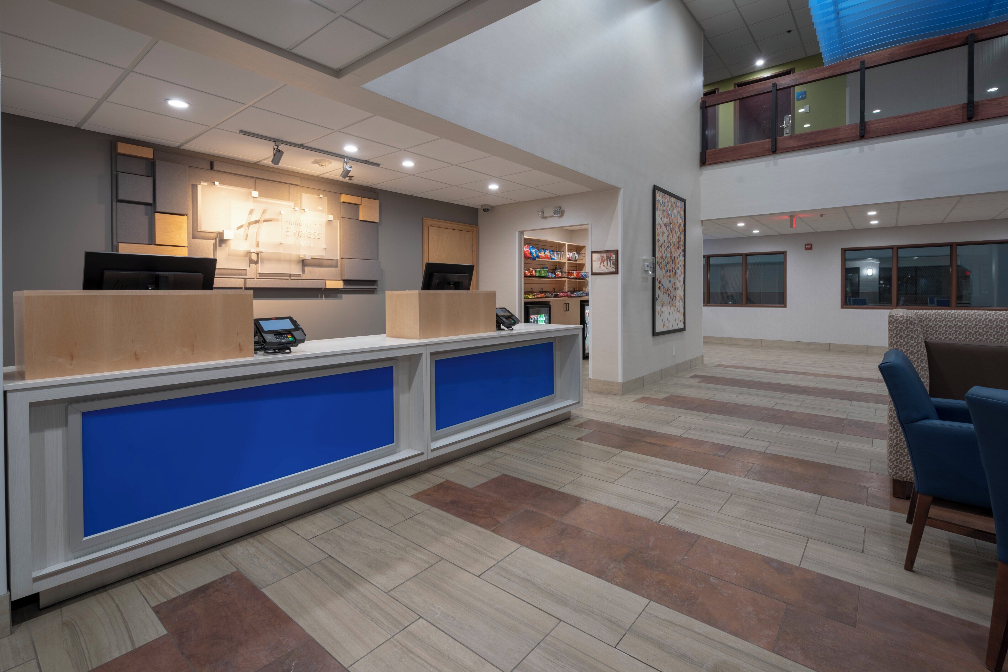 A hotel reception area with a counter, a digital kiosk, and a seating area with a blue chair.