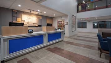 A hotel reception area with a counter, a digital kiosk, and a seating area with a blue chair.