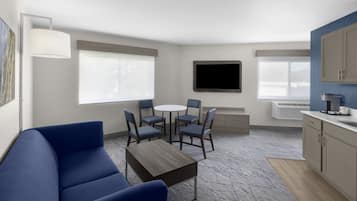 Una sala de estar moderna con un sofá azul, una mesa redonda y seis sillas. Hay un televisor de pantalla plana montado en la pared, una ventana con persianas y un área de cocina pequeña con armarios y una encimera.