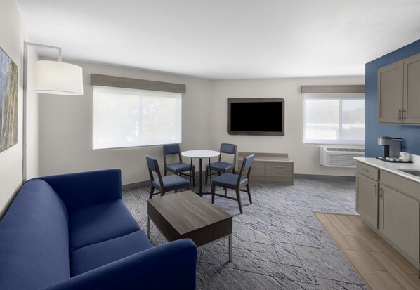 A modern living room with a blue sofa, a round table, and six chairs. There is a flat-screen TV mounted on the wall, a window with blinds, and a kitchenette area with cabinets and a countertop.