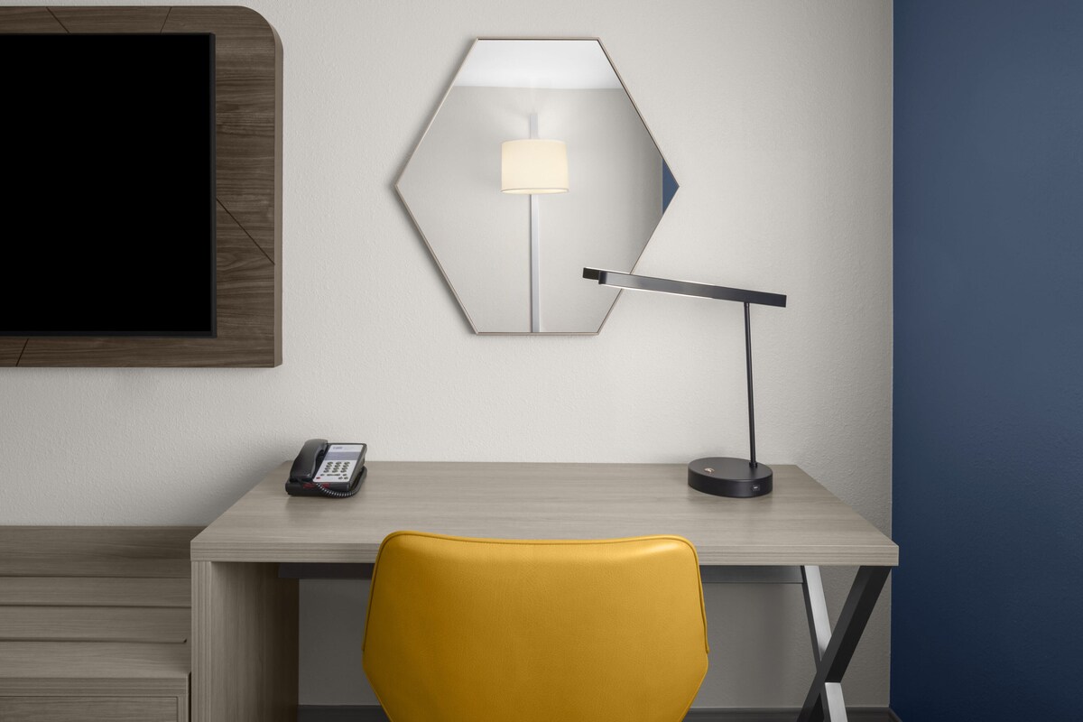Ein Hotelzimmer mit einem Holztisch, einem gelben Sessel, einem sechseckigen Spiegel, einer Lampe und einem Telefon.