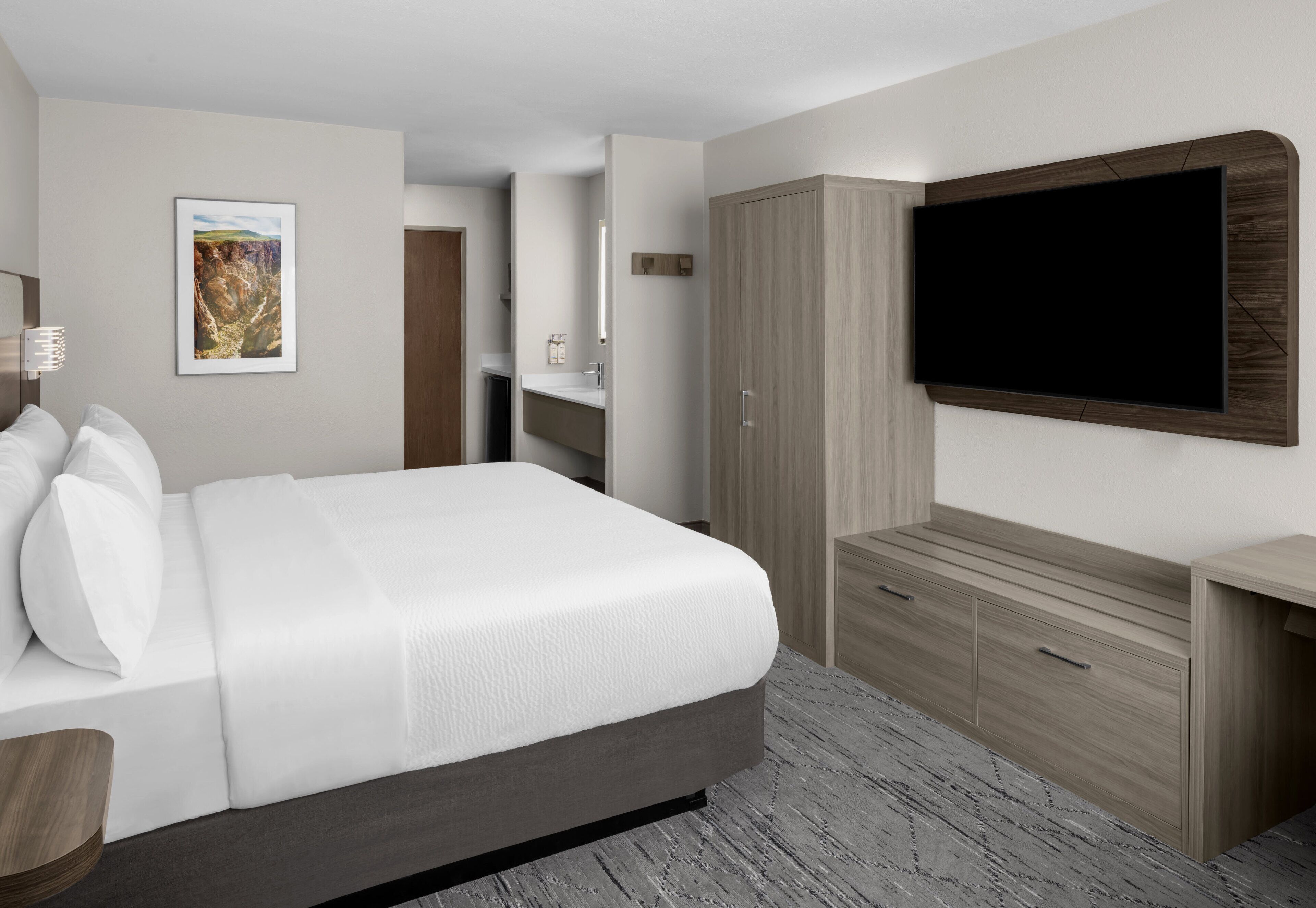 Ett modernt hotellrum med en stor säng, en platt-TV som är monterad på väggen, en garderob i trä och ett badrum som syns genom en öppen dörr.