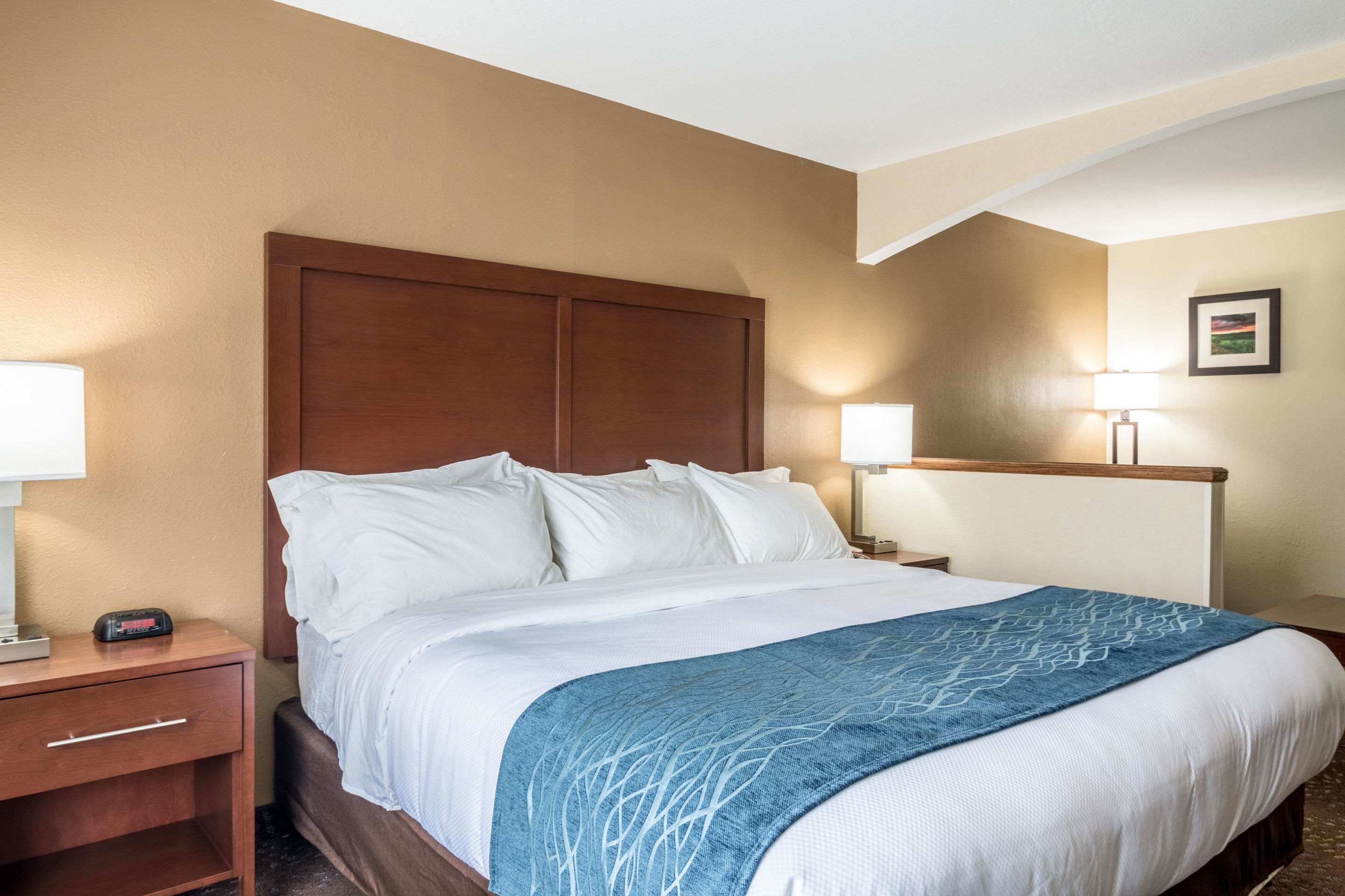 A hotel room with a large bed, wooden headboard, two bedside tables with lamps, and a nightstand with a clock.