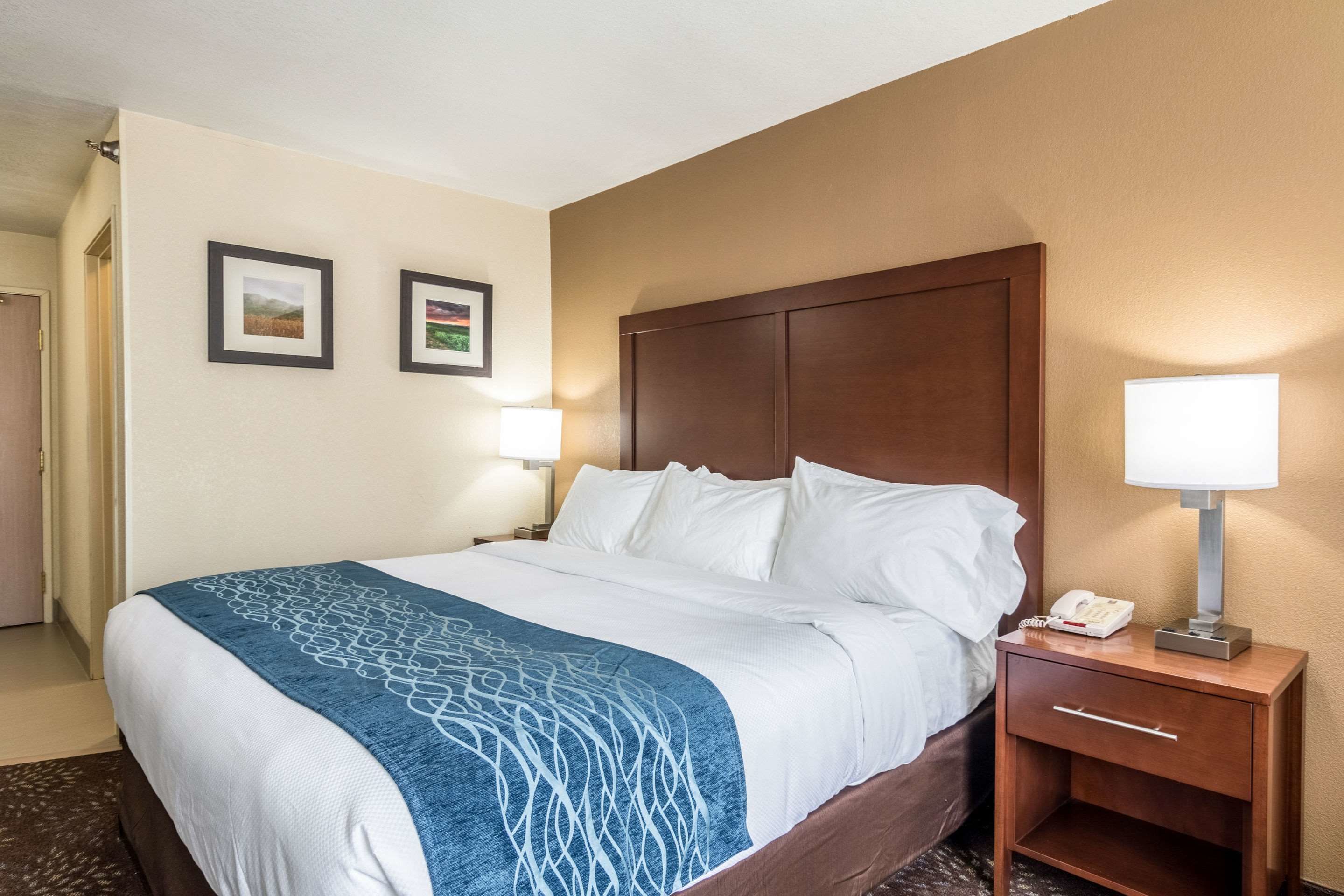 A hotel room with a large bed, a wooden headboard, two bedside lamps, a bedside telephone, and a small drawer unit.