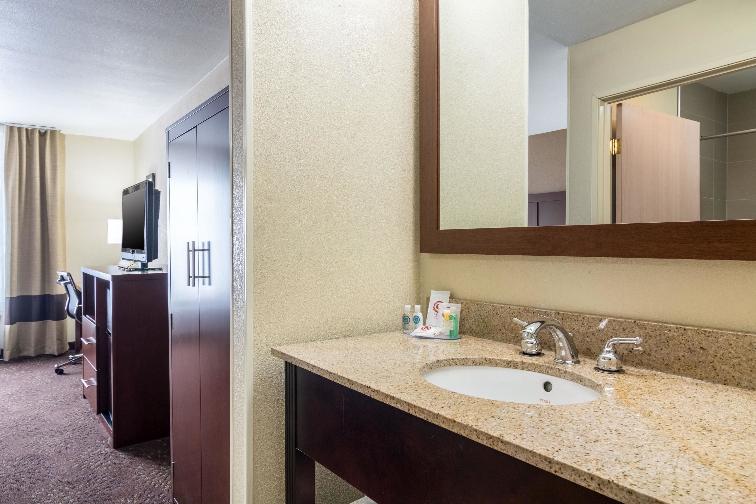 Un baño de hotel con lavamanos de encimera de granito, un espejo y un televisor al fondo.