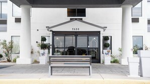 Entrada moderna de un edificio con el número "7520" sobre la puerta, un banco al frente y plantas en macetas a ambos lados.