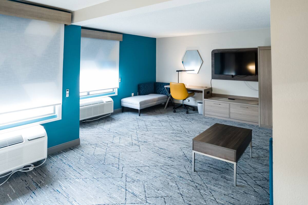 Ein modernes Zimmer mit blauer Akzentwand, einem grauen Sofa, einem gelben Sessel, einem Holztisch und einem Schreibtisch mit Lampe. Ein Fernseher ist an der Wand montiert.