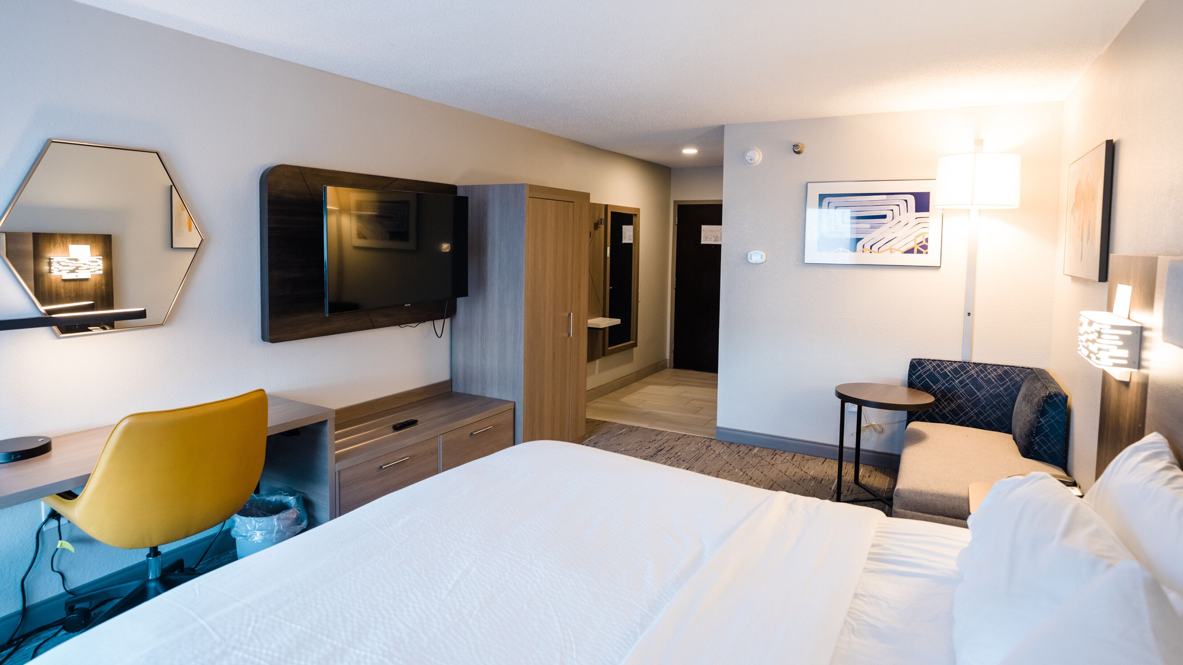 A hotel room with a bed, a desk with a chair, a TV, a wardrobe, a small table, and a lamp.