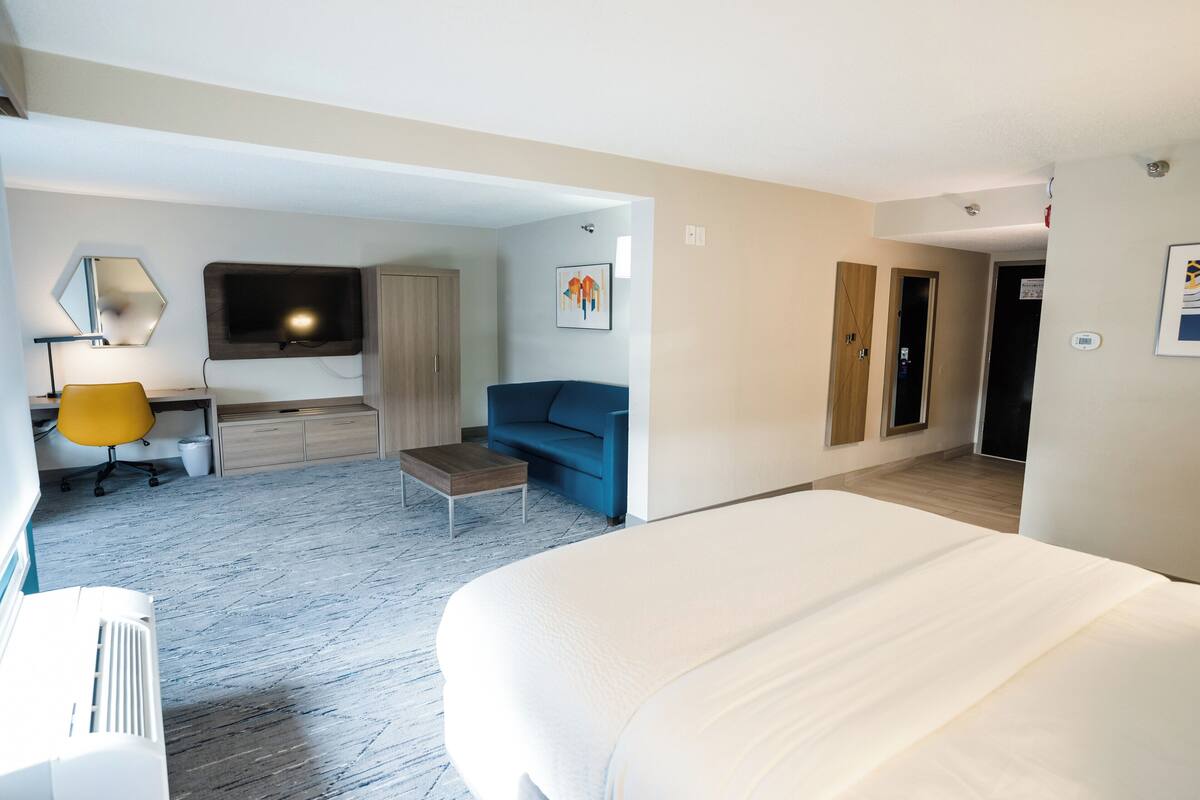 Ein Hotelzimmer mit einem Bett, einem Schreibtisch mit Stuhl, einer Couch, einem an der Wand montierten Fernseher und einer Tür zu einem weiteren Raum.