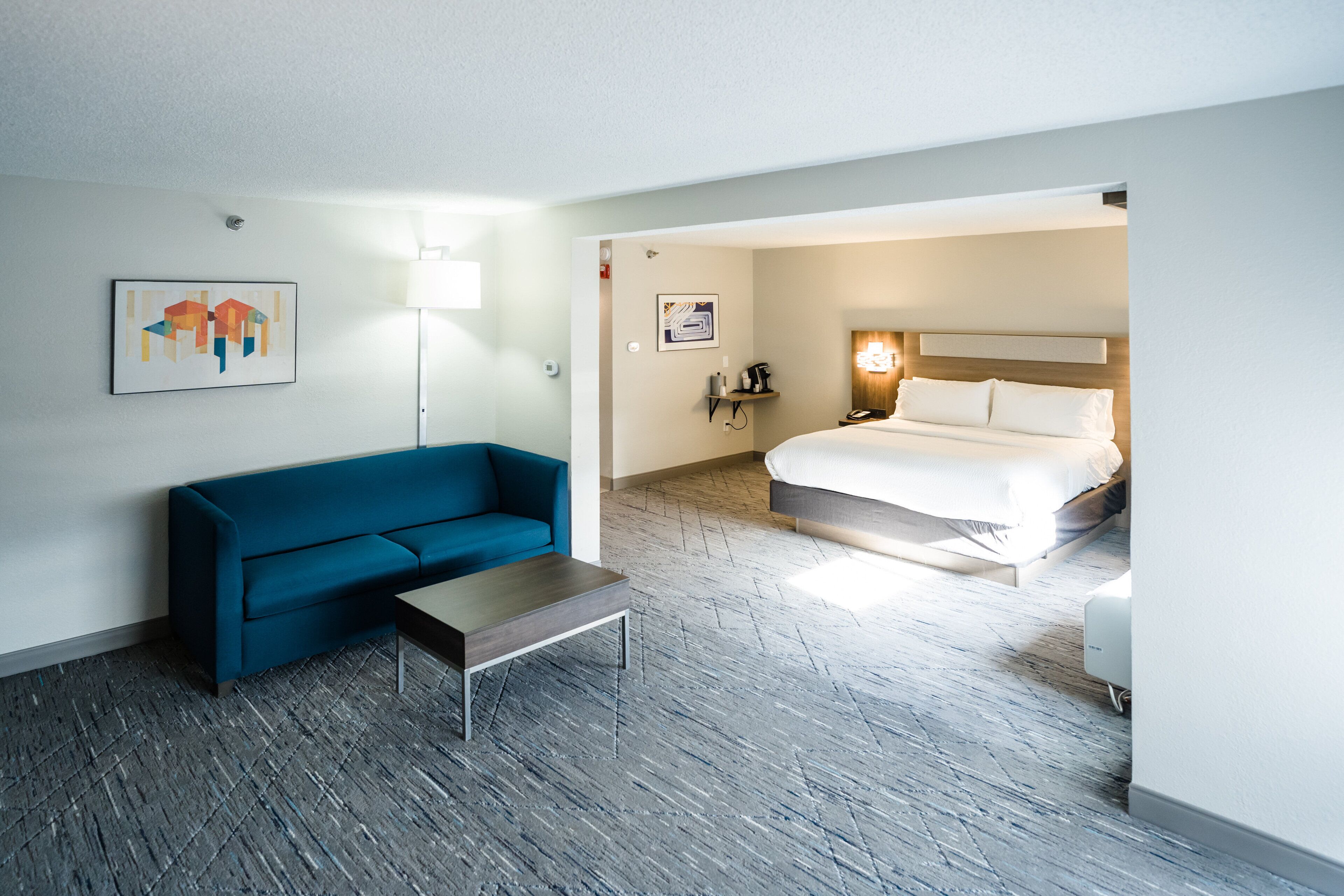 Habitación de hotel con un sofá azul, una mesita, una cama con ropa blanca, una lámpara y obras de arte en las paredes.