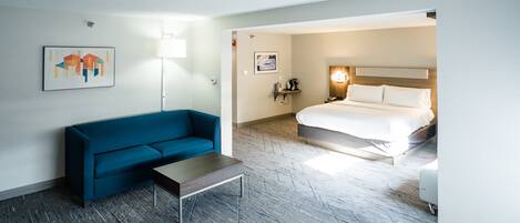 Habitación de hotel con un sofá azul, una mesita, una cama con ropa blanca, una lámpara y obras de arte en las paredes.