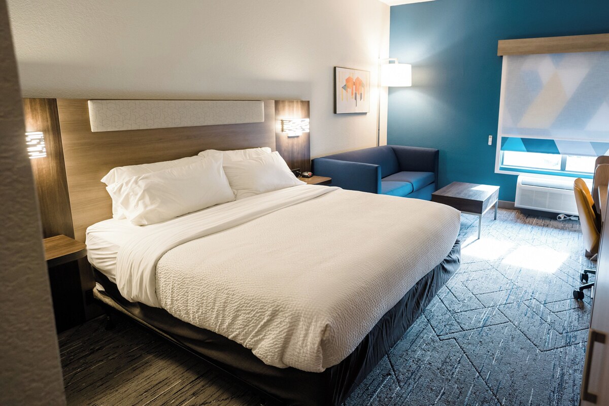 A hotel room with a large bed, a small sofa, a desk, and a window with blinds.