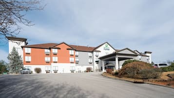 Situated on a street with a paved driveway, this hotel features a modern architectural design with a flat roof and a covered entrance area.