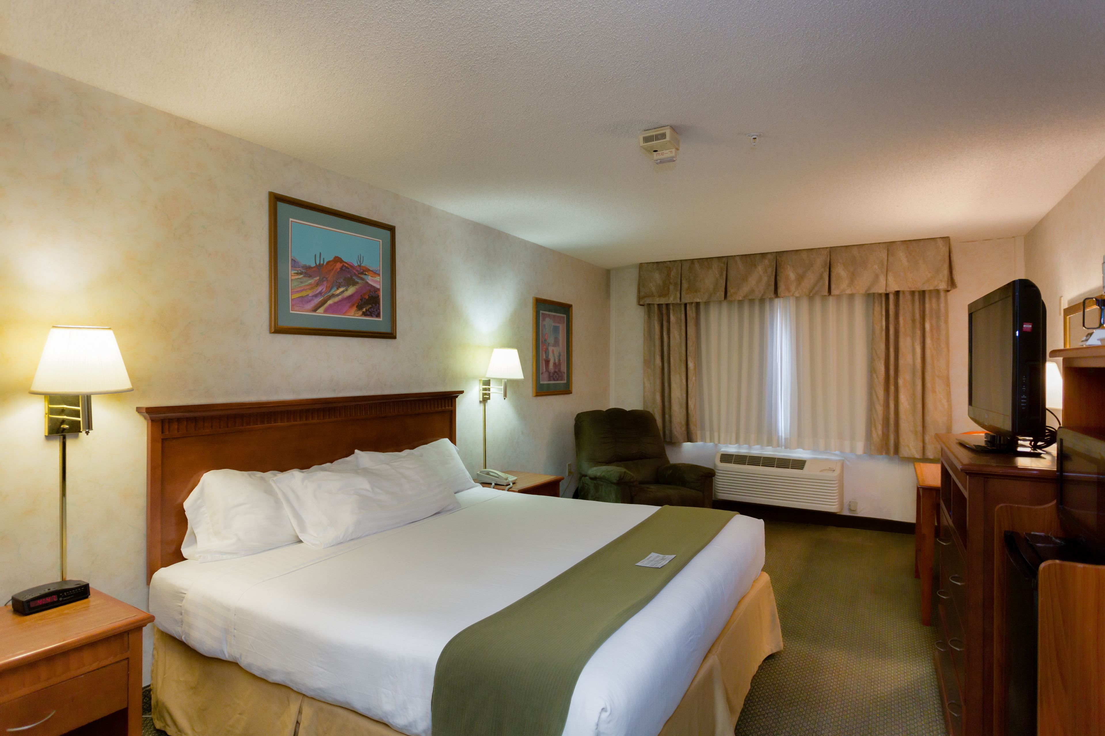 A hotel room with a large bed, a desk with a computer, a chair, a television, and a window with curtains.