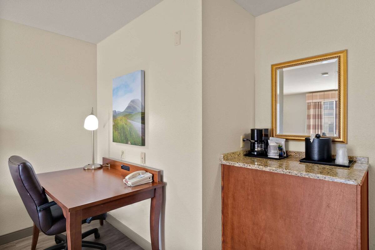 A hotel room with a wooden desk, a chair, a lamp, a painting, a coffee maker, and a mirror.