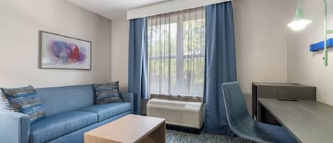 A hotel room with a sofa, a small table, a desk, a chair, a radiator, a window with curtains, and a wall-mounted lamp.