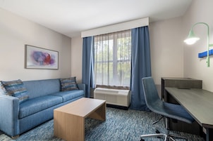 Habitación de hotel con sofá, una mesita, escritorio, silla, radiador, ventana con cortinas y una lámpara de pared.