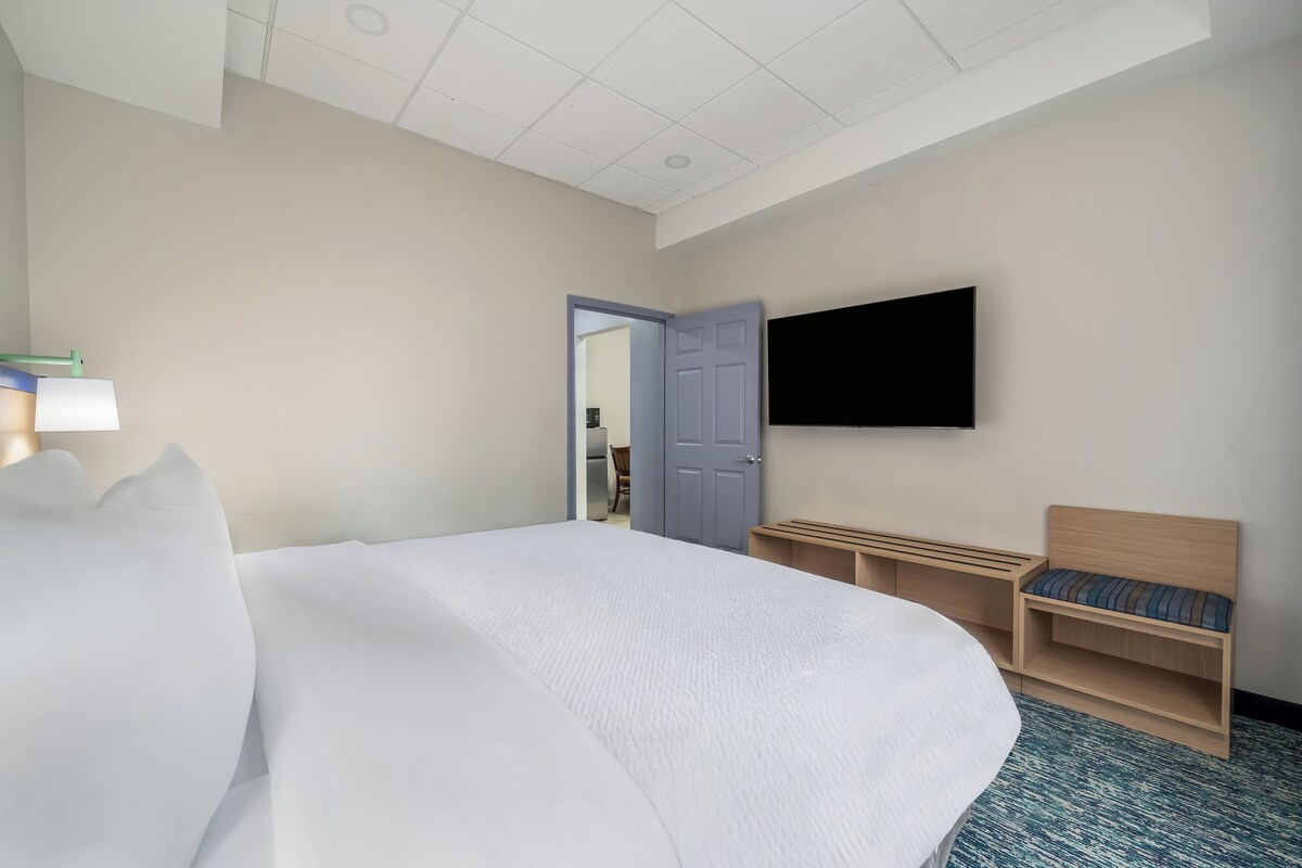 A hotel room with a large bed, a flat-screen TV mounted on the wall, a wooden bench with cushions, and a door leading to a bathroom.