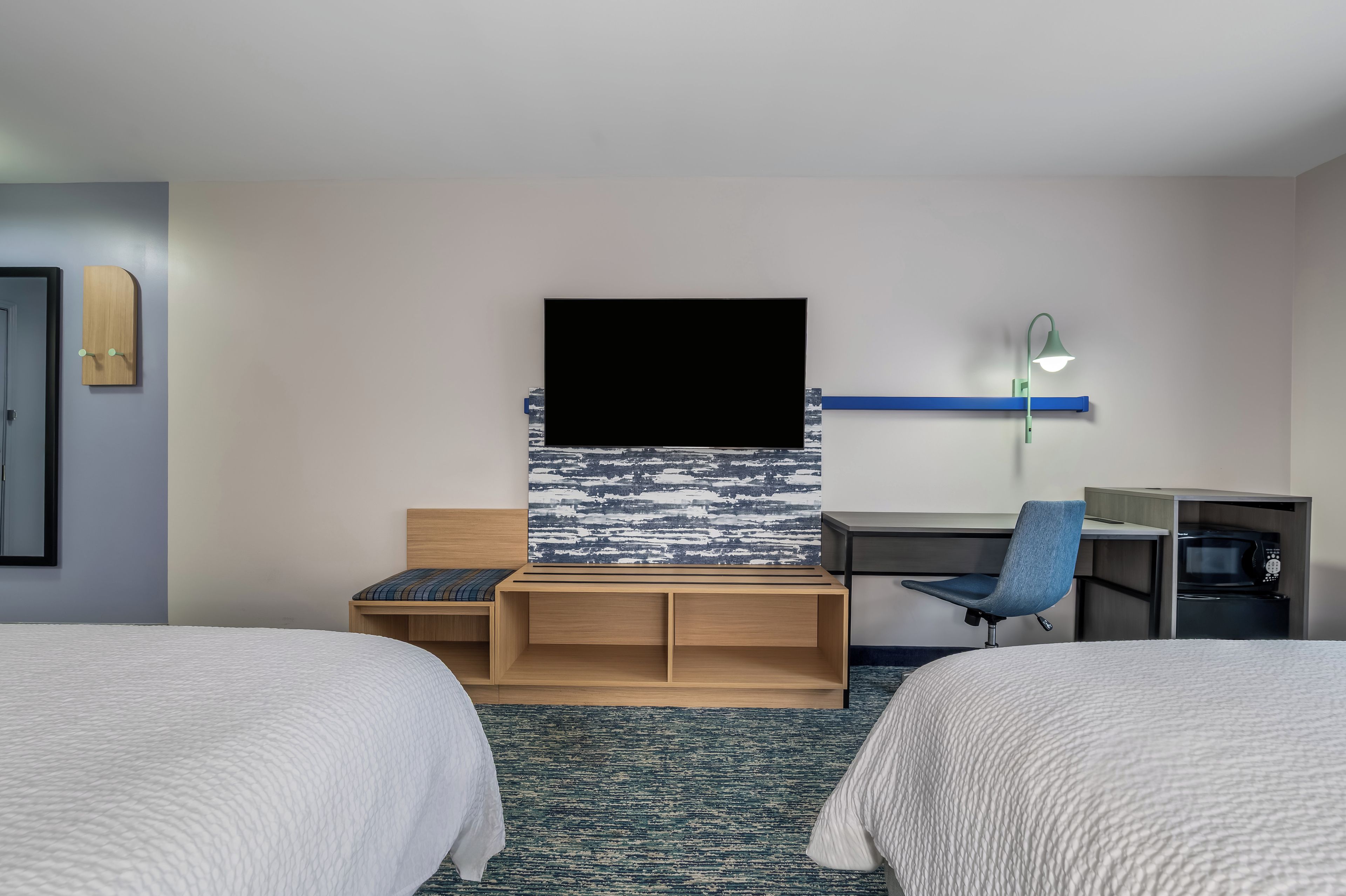 A hotel room with two beds, a desk with a chair, a flat-screen TV, a wall-mounted lamp, and a microwave.