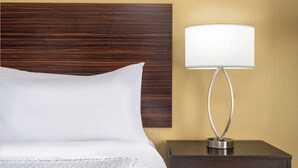 A neatly made bed with white linens, a wooden headboard, and a bedside table with a lamp. - Garner Hotel Birch Run Frankenmuth Area by IHG (Birch Run)