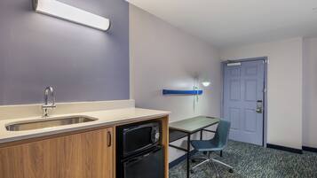 Una kitchenette con un lavamanos, lavavajillas y microondas. Hay un escritorio con una silla y una puerta al fondo.