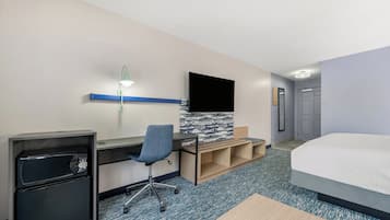 A hotel room with a bed, a desk with a computer, a chair, a small fridge, a TV, and a mirror.