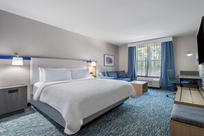 A hotel room with a large bed, a desk, a chair, a sofa, and a window with curtains. - Garner Hotel Birch Run Frankenmuth Area by IHG (Birch Run)