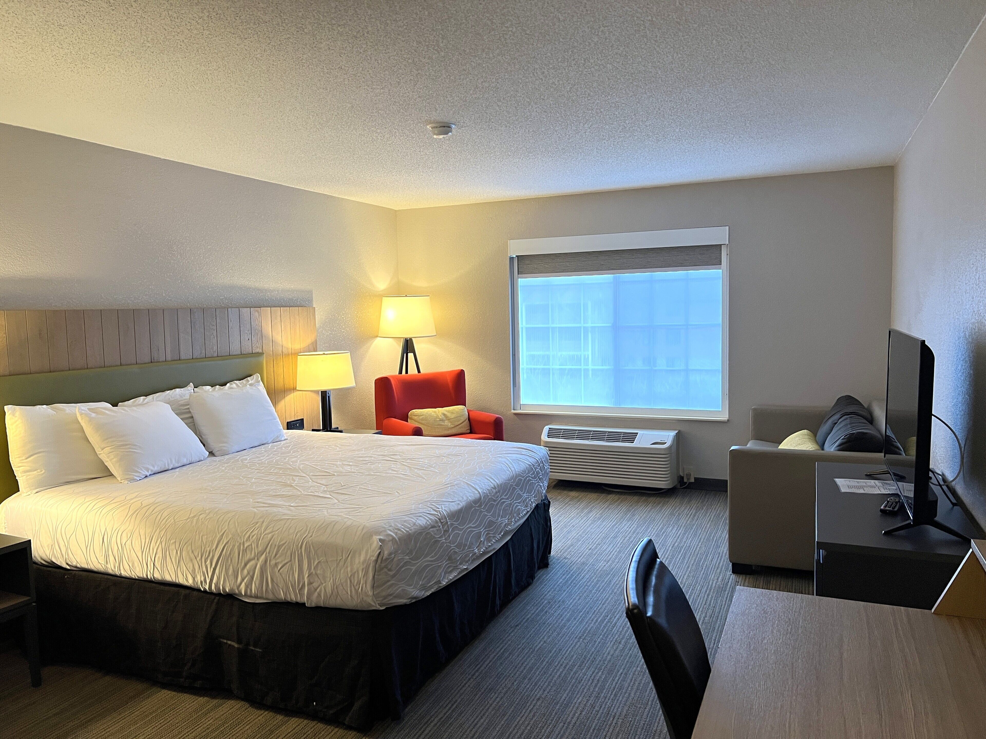 A hotel room with a large bed, a desk with a computer, a chair, a red armchair, a lamp, and a window with blinds.