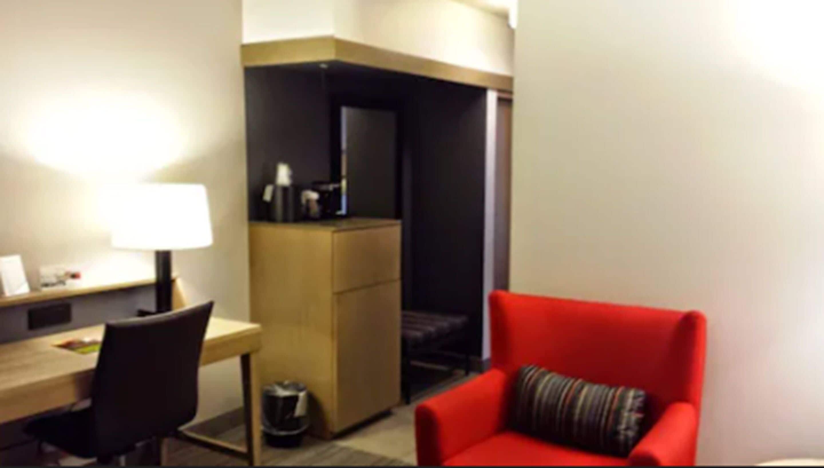A hotel room with a desk, chair, and a red armchair.