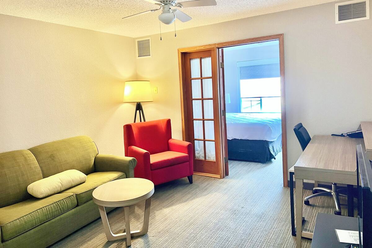 Une chambre d’hôtel avec un canapé vert, un fauteuil rouge, une petite table ronde, un ventilateur de plafond, un lit près d’une fenêtre et un bureau avec une chaise.