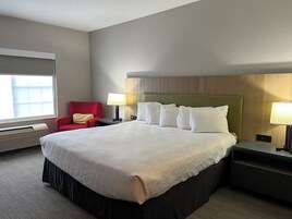 Habitación de hotel con una cama grande, un sillón rojo, dos mesitas de noche con lámparas, una ventana con persianas y un cabecero de madera.