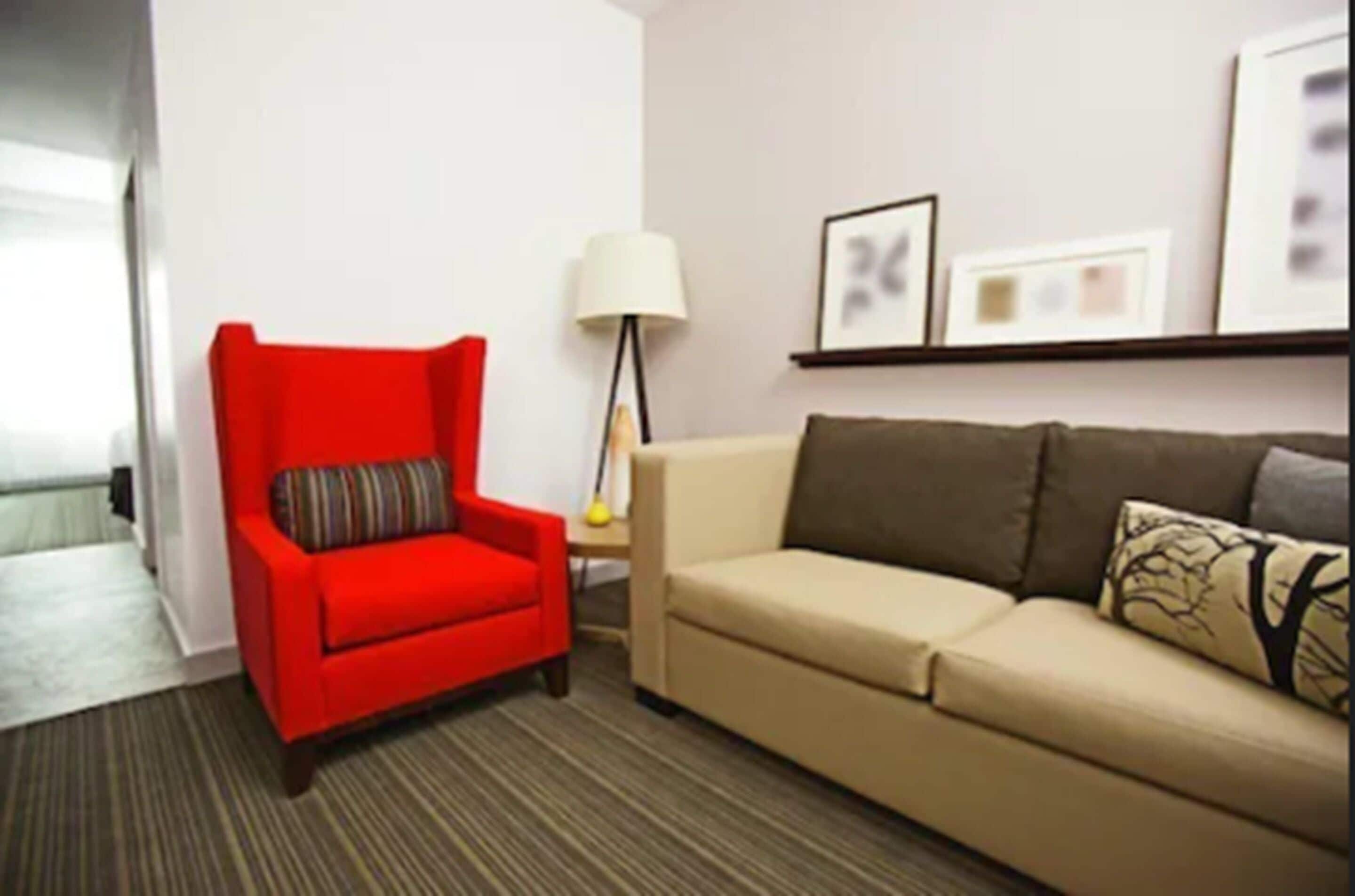 A room with a red armchair, a beige sofa, a lamp, and framed pictures on the wall.