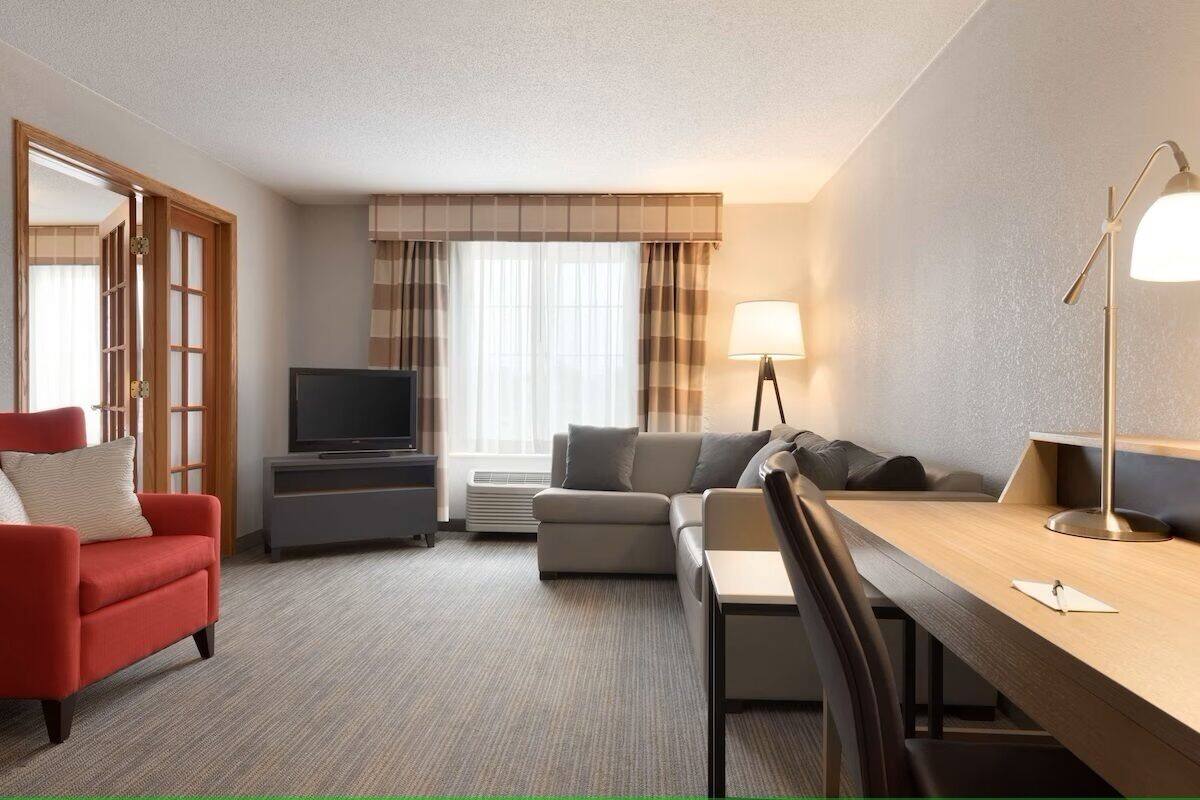 Une chambre d’hôtel avec un fauteuil rouge, un canapé, une télévision, un bureau avec une lampe et une grande fenêtre avec des rideaux.