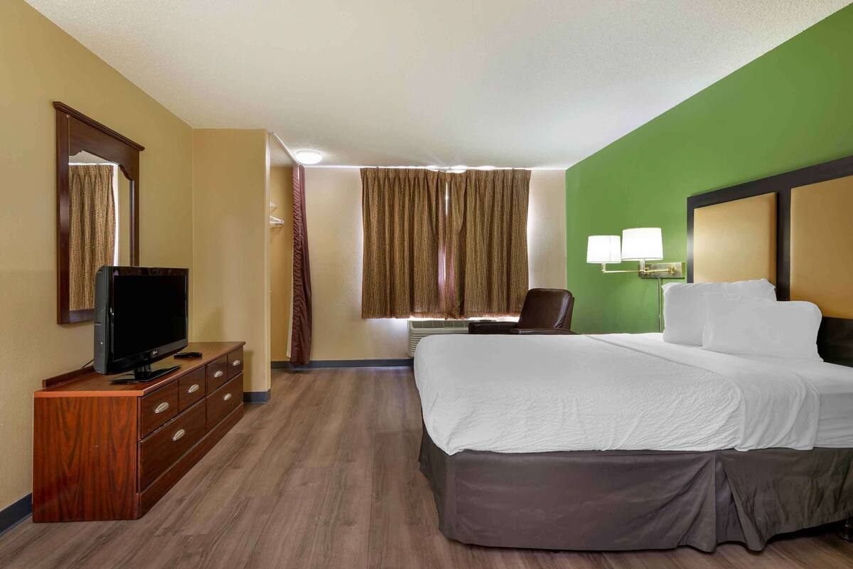 Habitación de hotel con una cama grande, un televisor sobre una cómoda de madera, un escritorio con una silla y una ventana con cortinas.