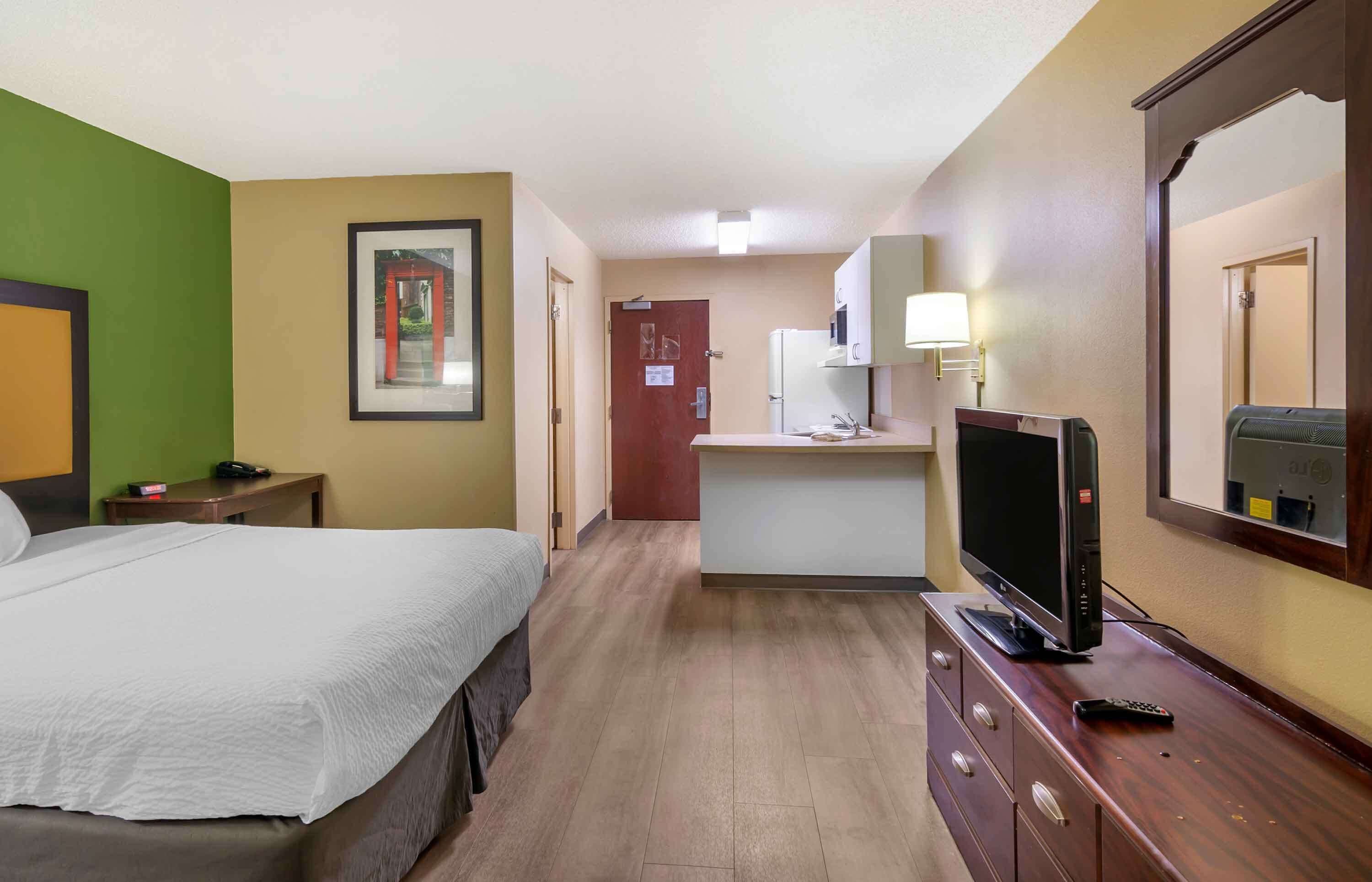 Quarto de hotel com cama, televisão, escrivaninha e cozinha compacta.