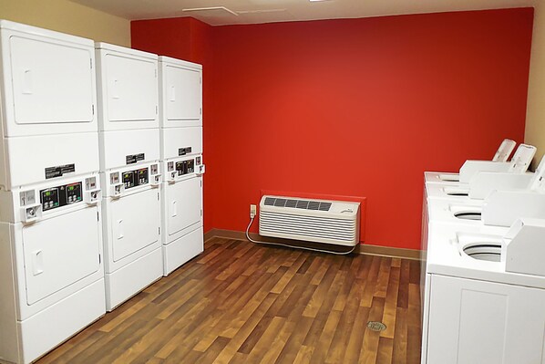 A laundry room with multiple white dryers, a wall-mounted air conditioner, and a wooden floor. - Extended Stay America Suites Dallas Frankford Road (Dallas)