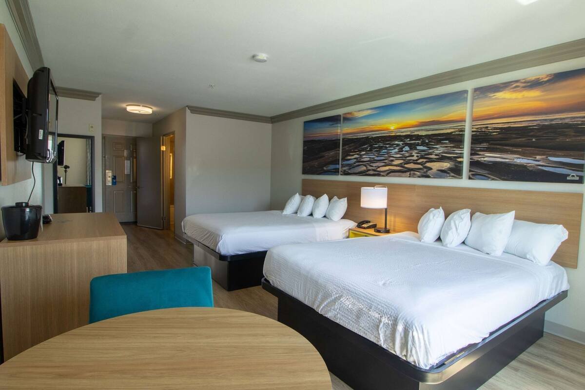 A hotel room with two beds, a desk, a chair, a TV, and a painting of a sunset.