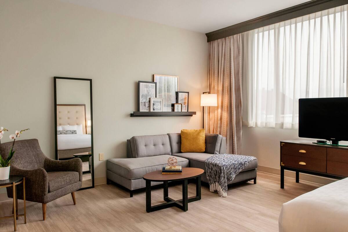A modern hotel room with a grey sofa, a small round table, a TV on a cabinet, a large mirror, and a window with sheer curtains.