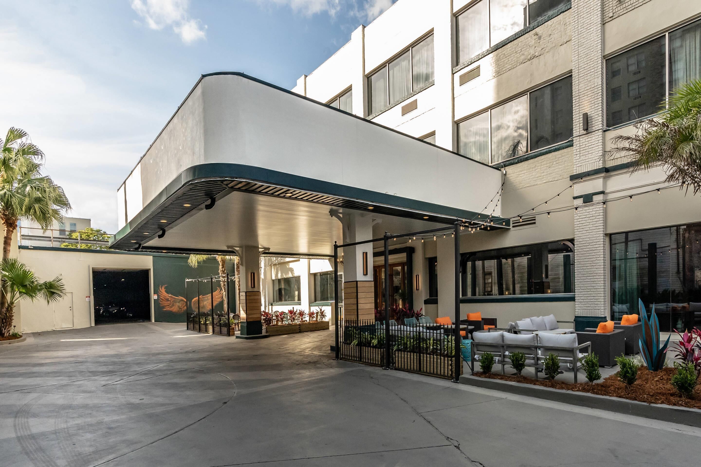 Een moderne hotelentree met een overdekt terras, een palmboom en een muurschildering.