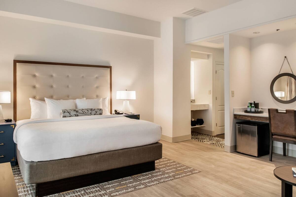 A hotel room with a large bed, bedside lamps, a small desk with a chair, and a mirror on the wall.
