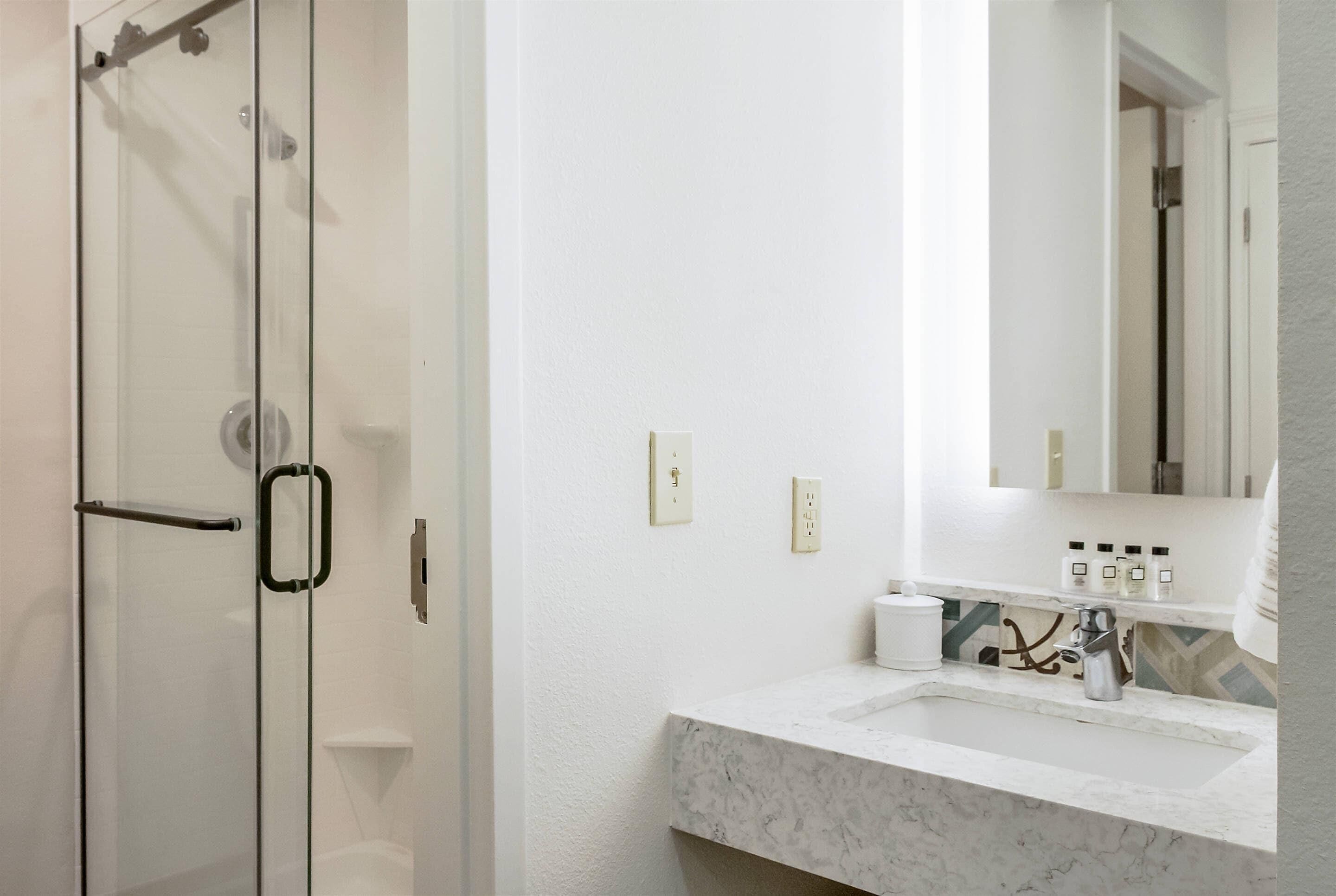 Une salle de bain dotée d’une douche à parois de verre, d’un lavabo blanc avec un plan de toilette en marbre et d’un miroir au-dessus du lavabo.