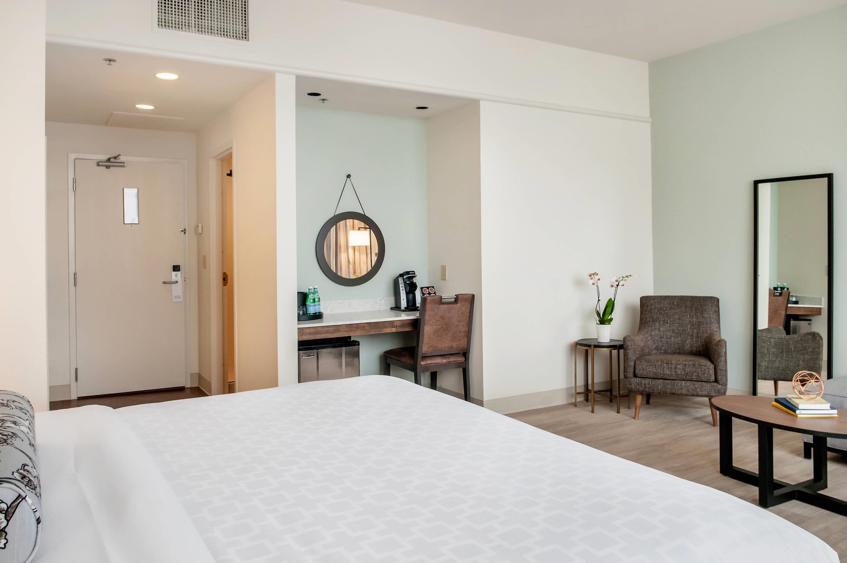 Habitación de hotel con una cama grande, un escritorio con silla, una mesita, un espejo y una planta en maceta.
