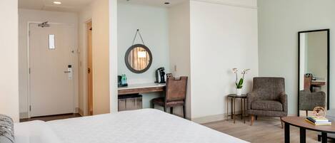 Habitación de hotel con una cama grande, un escritorio con silla, una mesita, un espejo y una planta en maceta.
