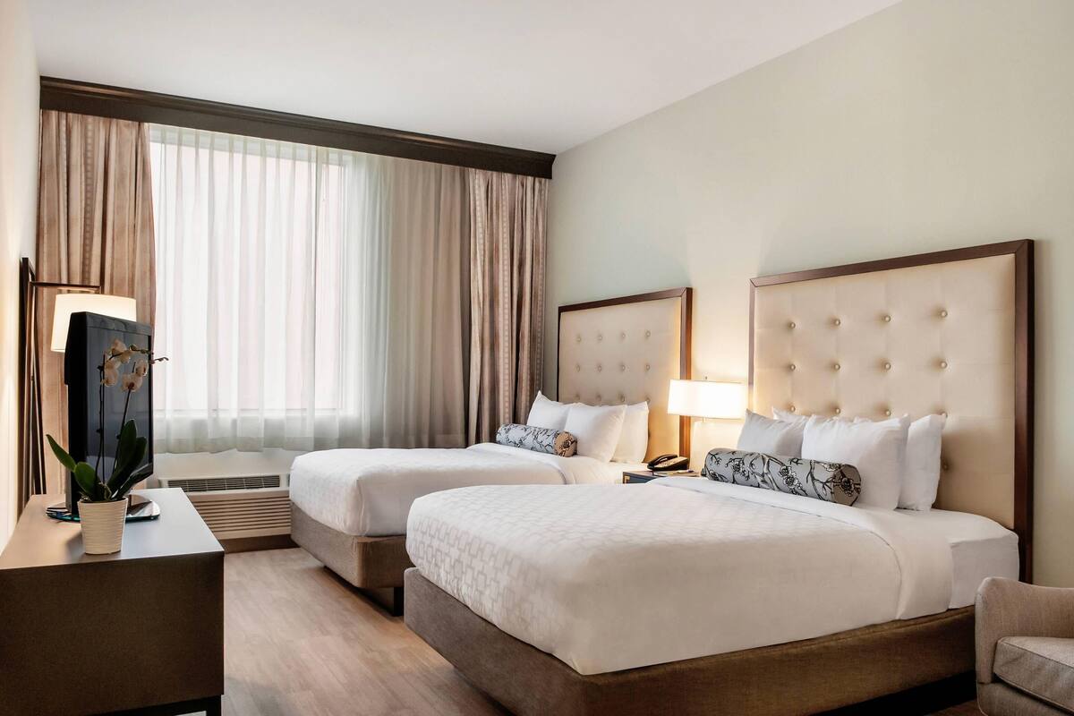 A hotel room with two beds, a desk with a lamp, and a large window with curtains.