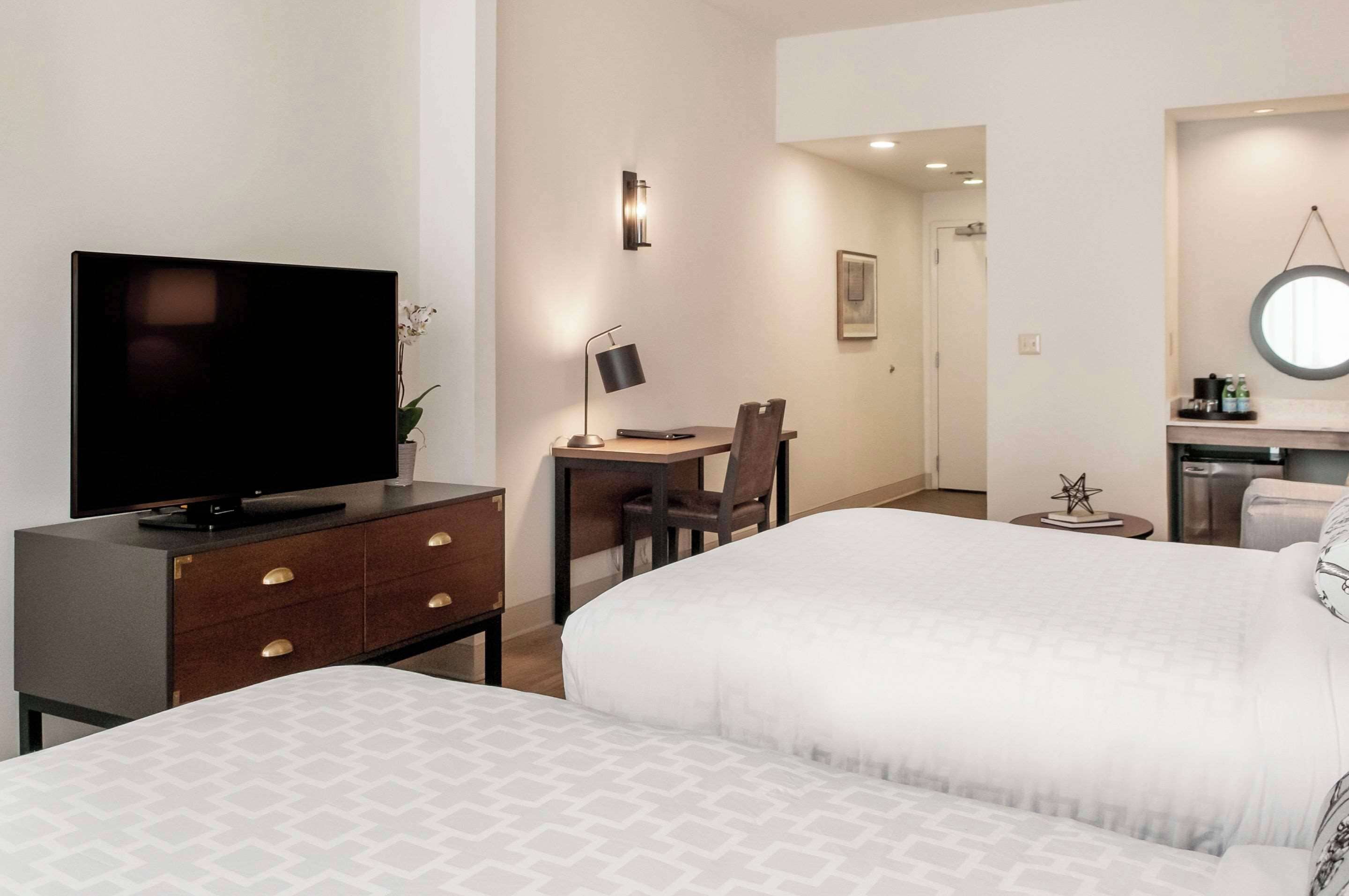 A hotel room with a bed, a desk with a chair, a television, and a small table with a lamp.