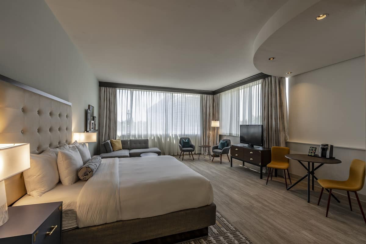 A hotel room with a large bed, a desk, two chairs, a TV, and a window with curtains.