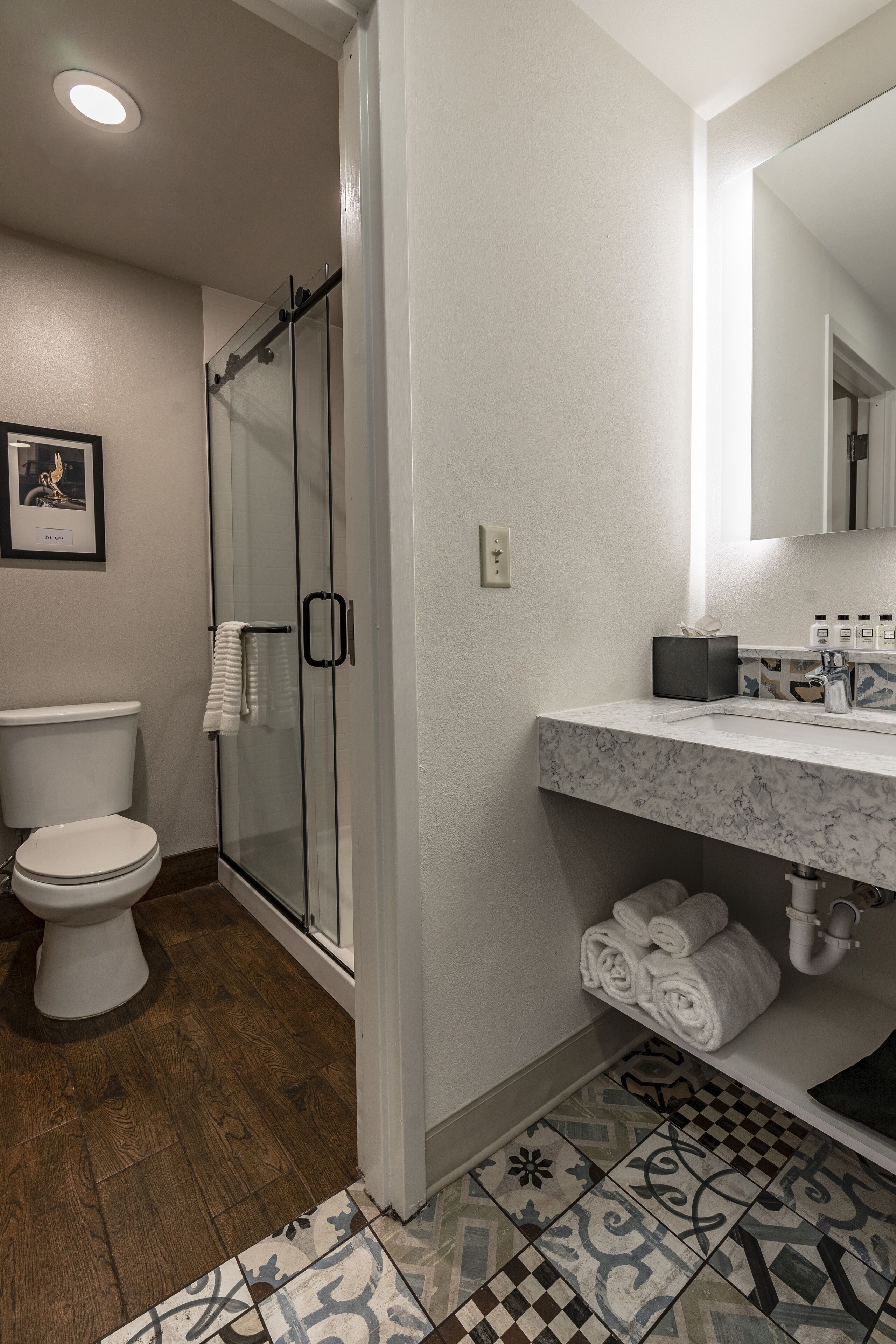 A bathroom with a glass shower enclosure, a white toilet, a marble sink with toiletries, and patterned tiles.