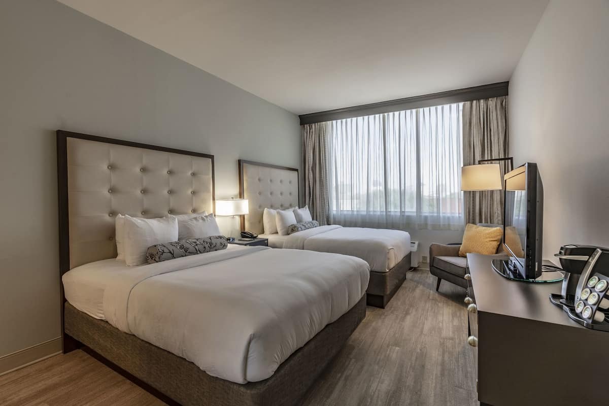 A hotel room with two beds, a desk with a computer, a chair, and a large window with curtains.